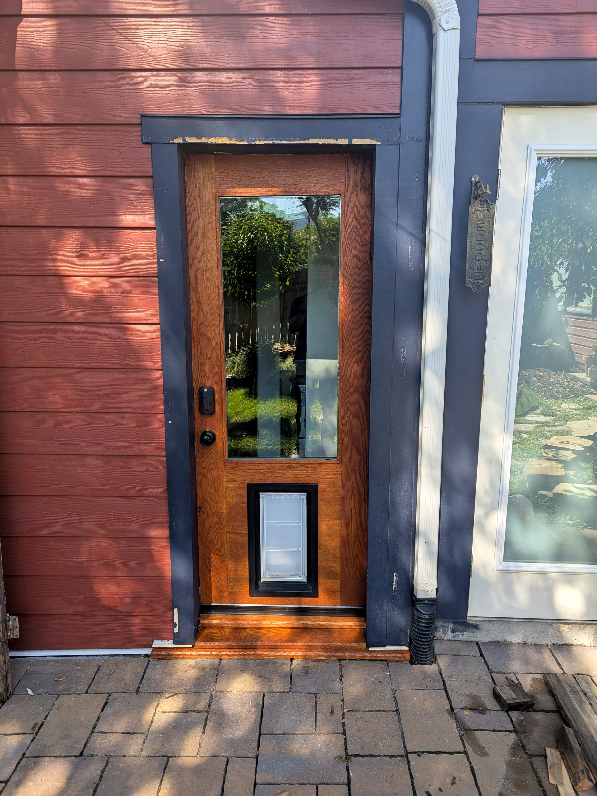 The finished white oak back door installed on the red siding — single pane of insulated glass reflecting the green backyard, oversized dog door, matte black hardware