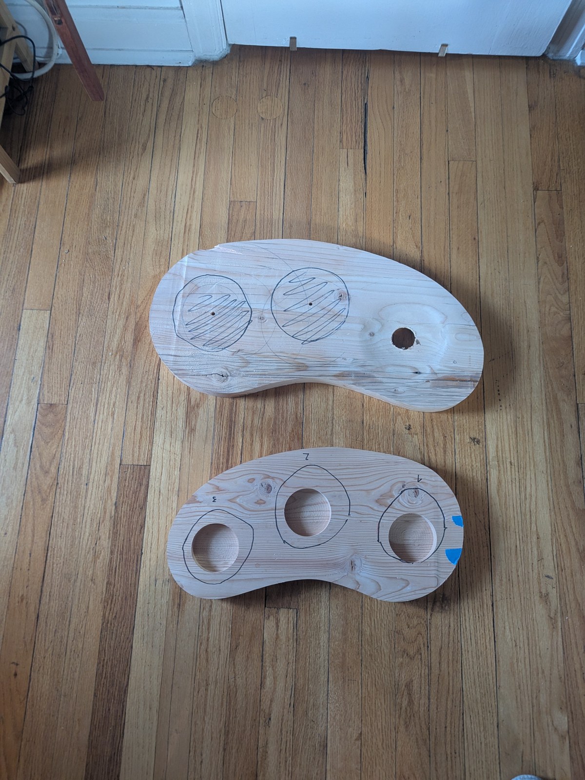 Two softwood prototype bases on the floor: one with machined round pockets, one marked out for hand-carved recesses